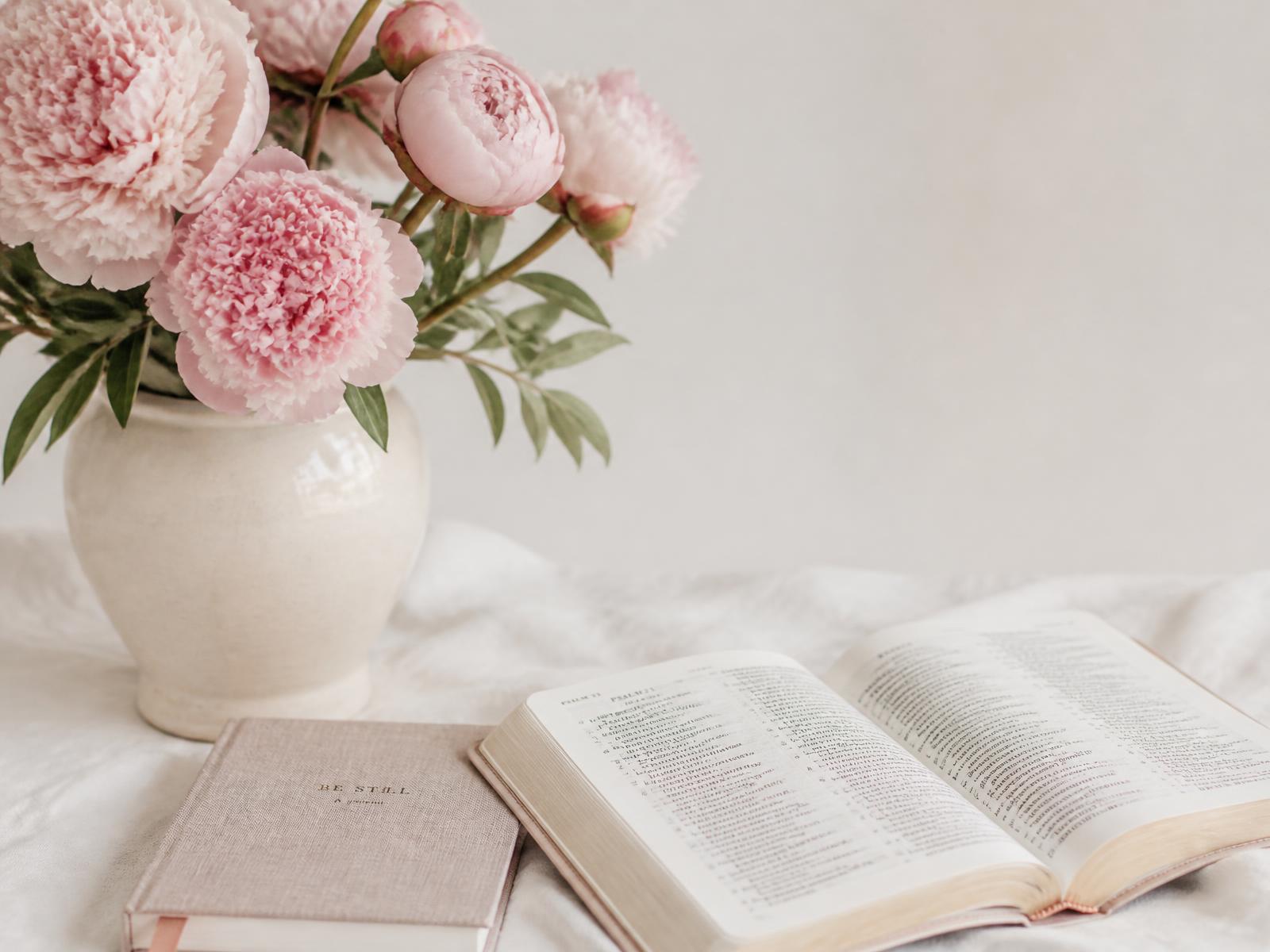 Pink peonies in a cream ceramic vase beside an open Bible and a linen notebook